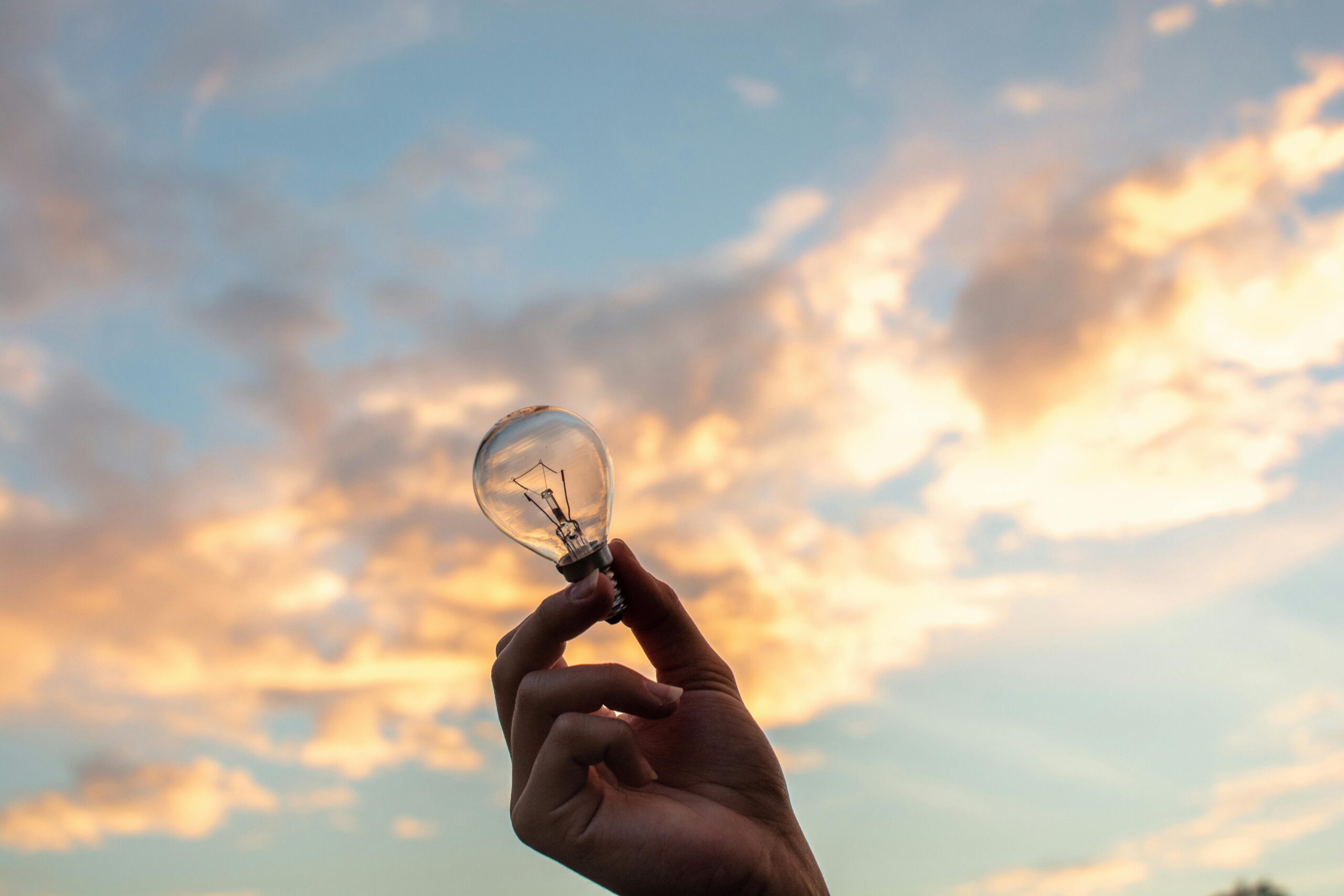 Someone's hand holding a lightbulb outside, against a setting sky.