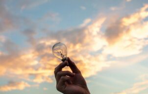 Someone's hand holding a lightbulb outside, against a setting sky.